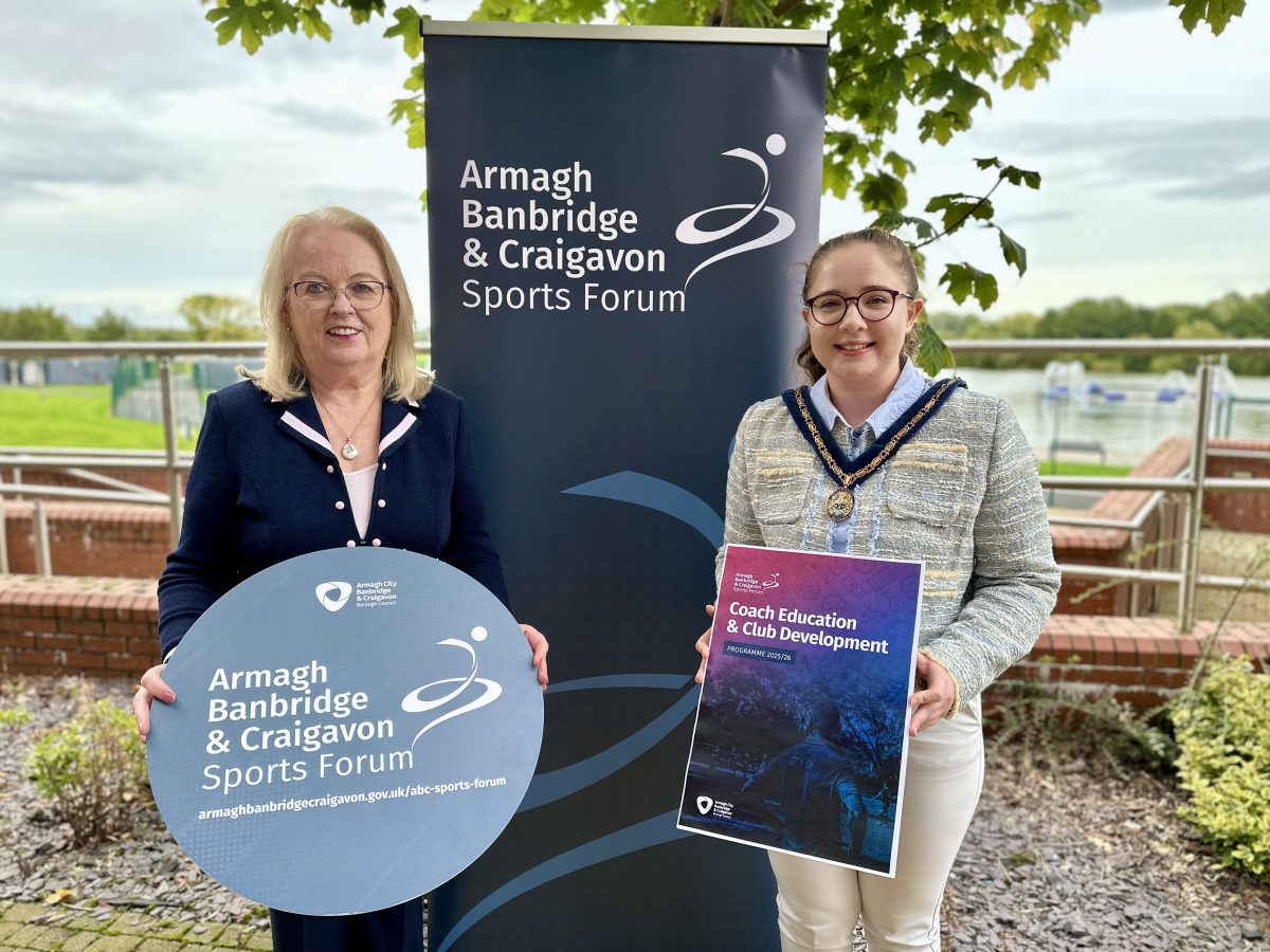Two women standing in front of the ABC Sports Forum banner. Each woman is holding a sign to promote the Coach Education and Club Development programme.