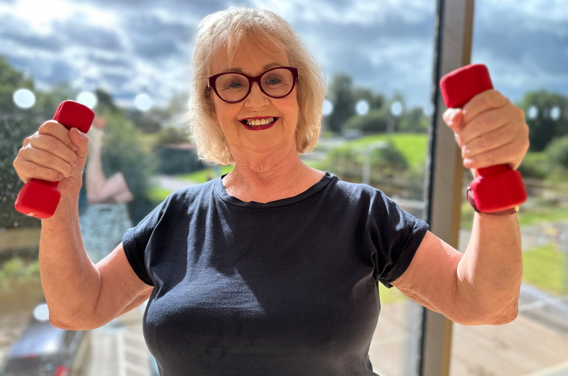 A older lady is smiling and holding two light dumbells.