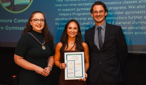Three people standing in front of a large screen an the NI Gymnastics Awards.