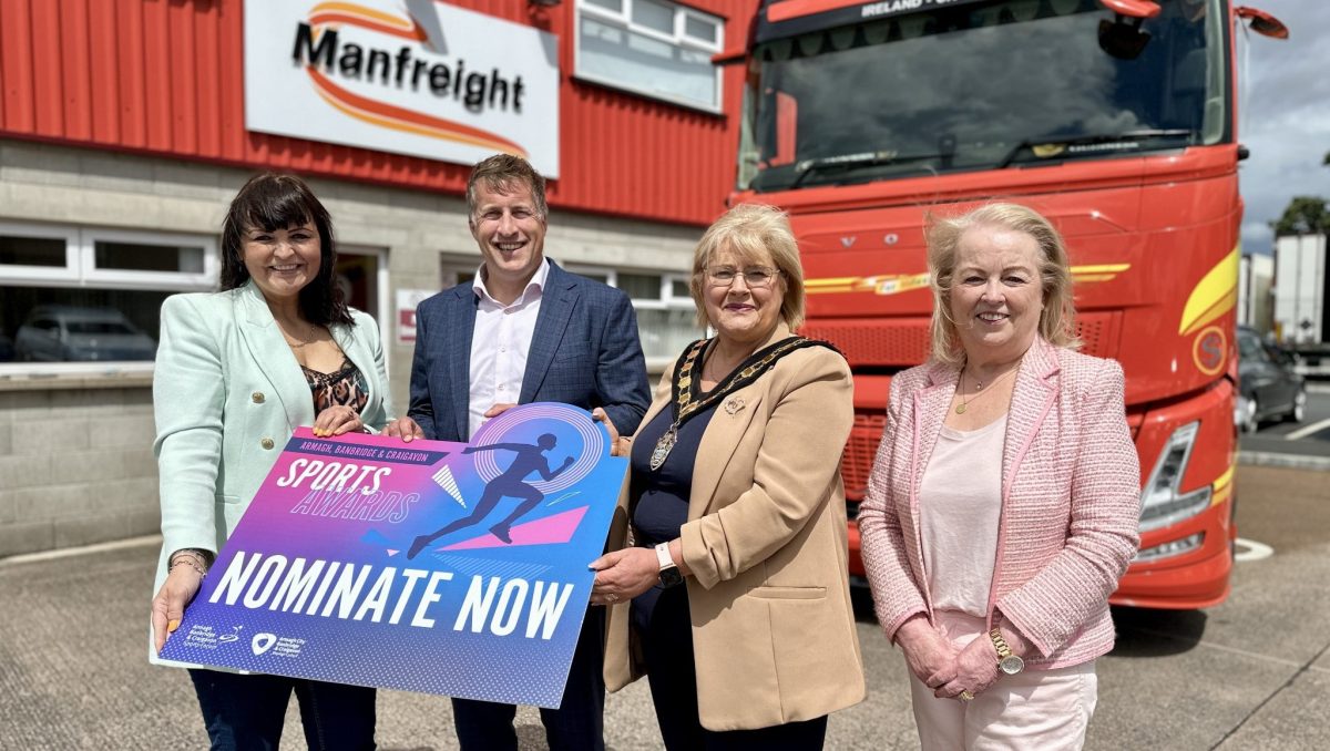 Four people standing in front of a red and yellow lorry with Manfreight branding and holding a banner to promore the ABC Junior Sports Awards.